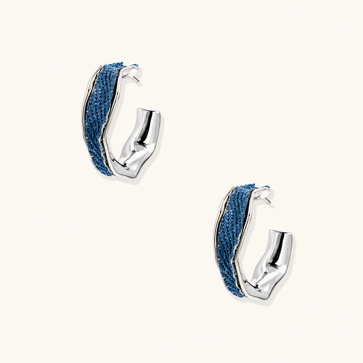 Silver huggie hoops with blue denim fabric inlay strip and polished white gold plated finish on cream background.