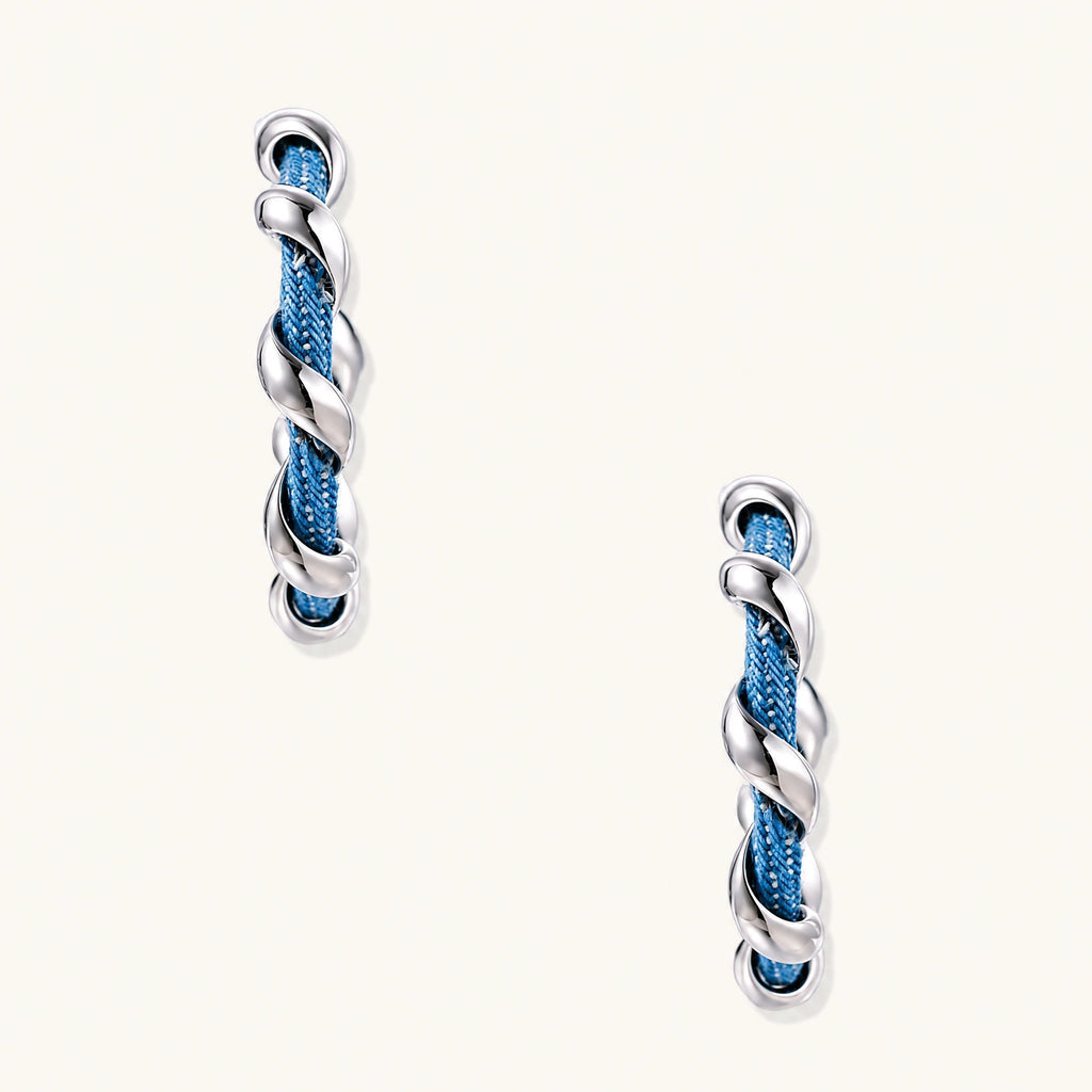 White gold-plated hoops intertwined with blue denim fabric in a twisted rope design on cream background.