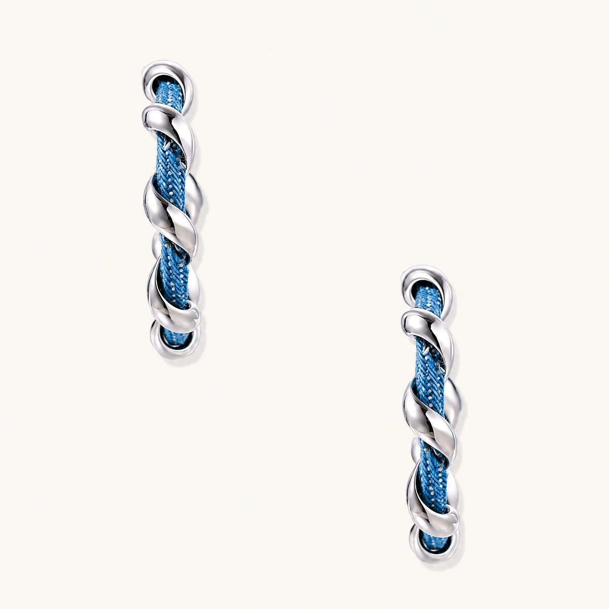 White gold-plated hoops intertwined with blue denim fabric in a twisted rope design on cream background.