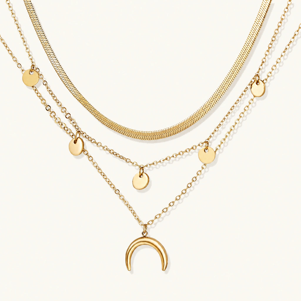 Four-layer gold necklace with herringbone chain, disc charms, and crescent moon pendant on cream background