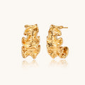 Pair of gold huggie hoops with sculpted wave texture and dimensional ripples on neutral background.