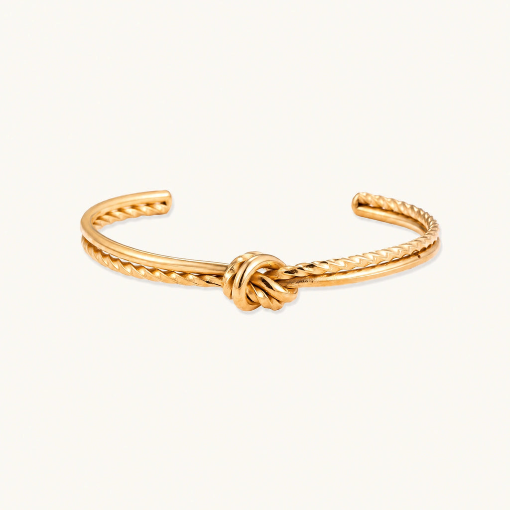 Gold cuff bracelet with twisted rope texture and decorative knot centerpiece on cream background.